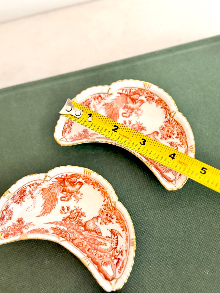 Two small decorative ceramic dishes with red toile-style prints and gold trim, shown on a dark green surface with a yellow measuring tape laid across one dish to indicate size.
